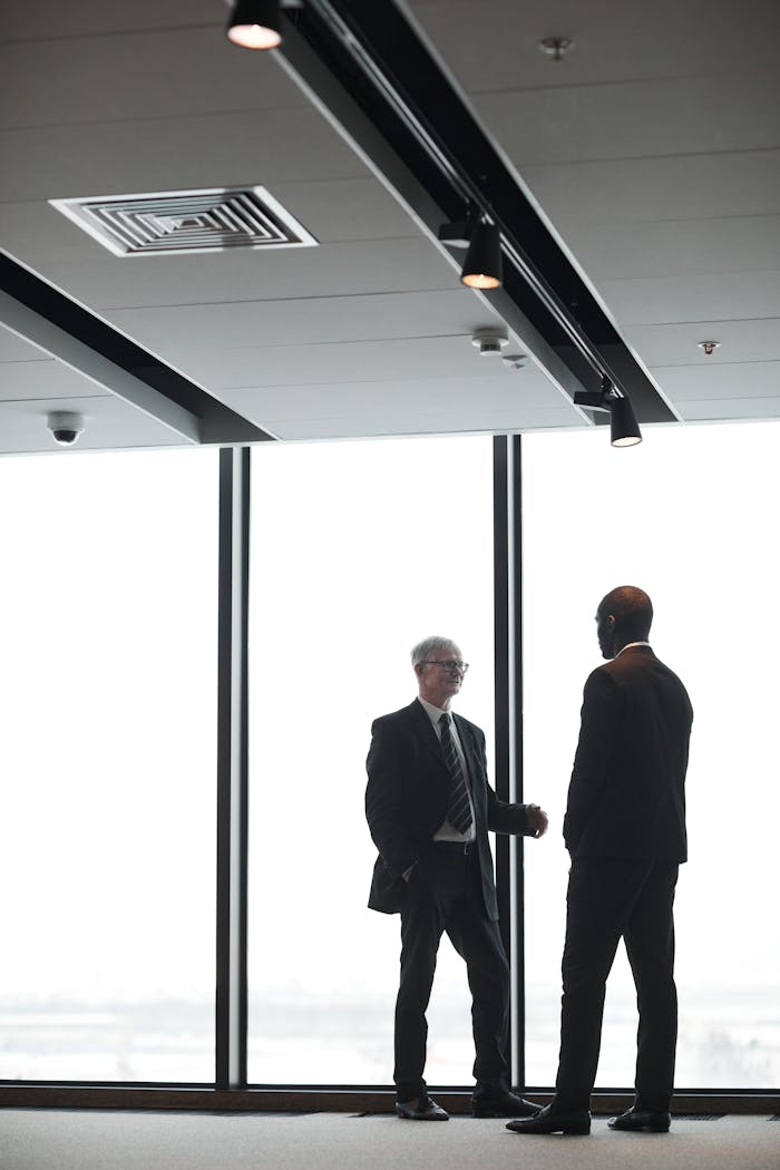 services-04 Two corporate executives in formal attire discuss business indoors, silhouetted against large windows.